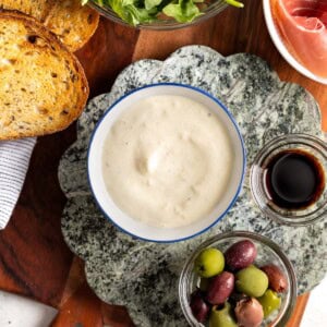 A bowl of lemon aioli surrounded by toasted bread, prosciutto, arugula, olives and balsamic.