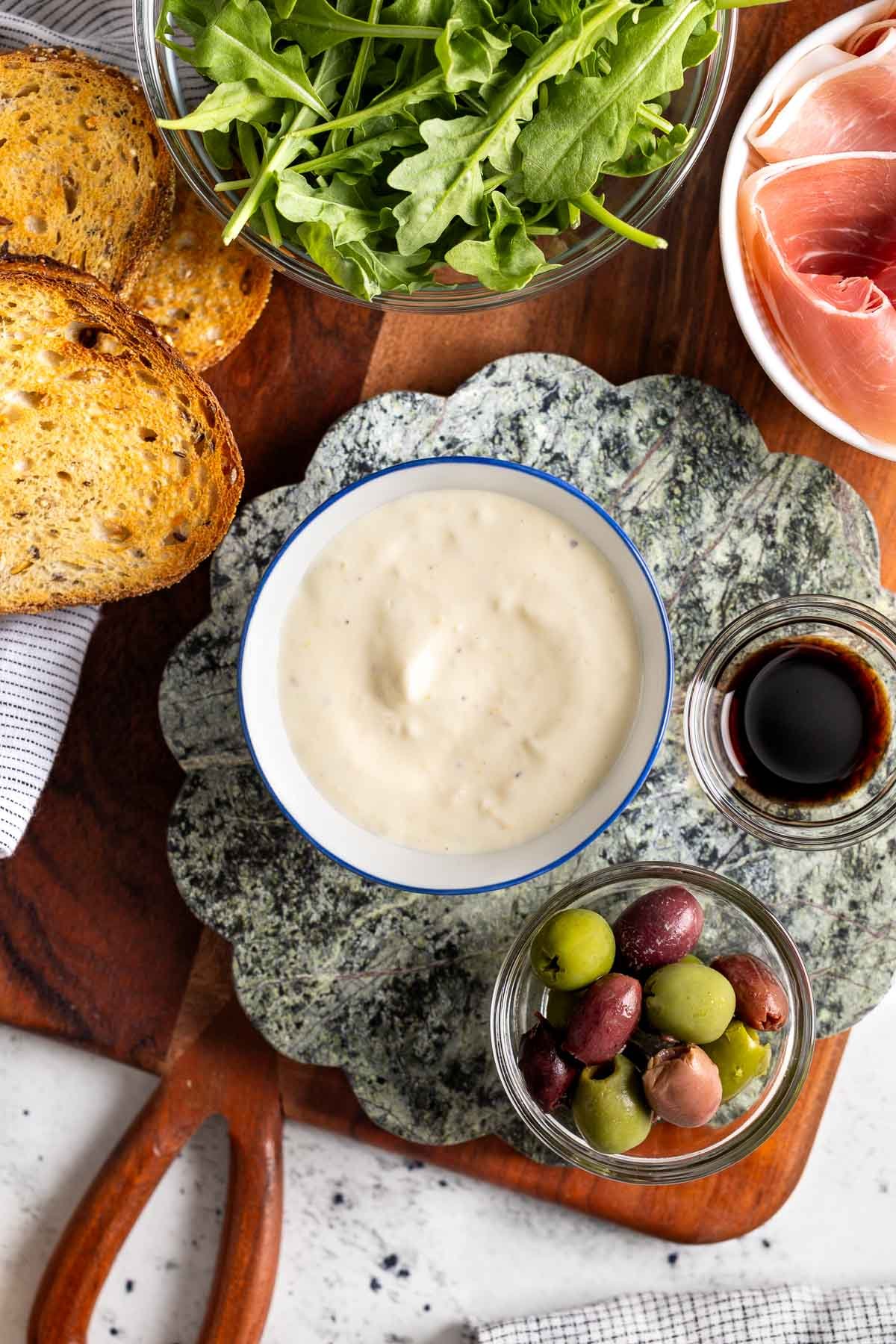 A bowl of lemon aioli surrounded by toasted bread, prosciutto, arugula, olives and balsamic.