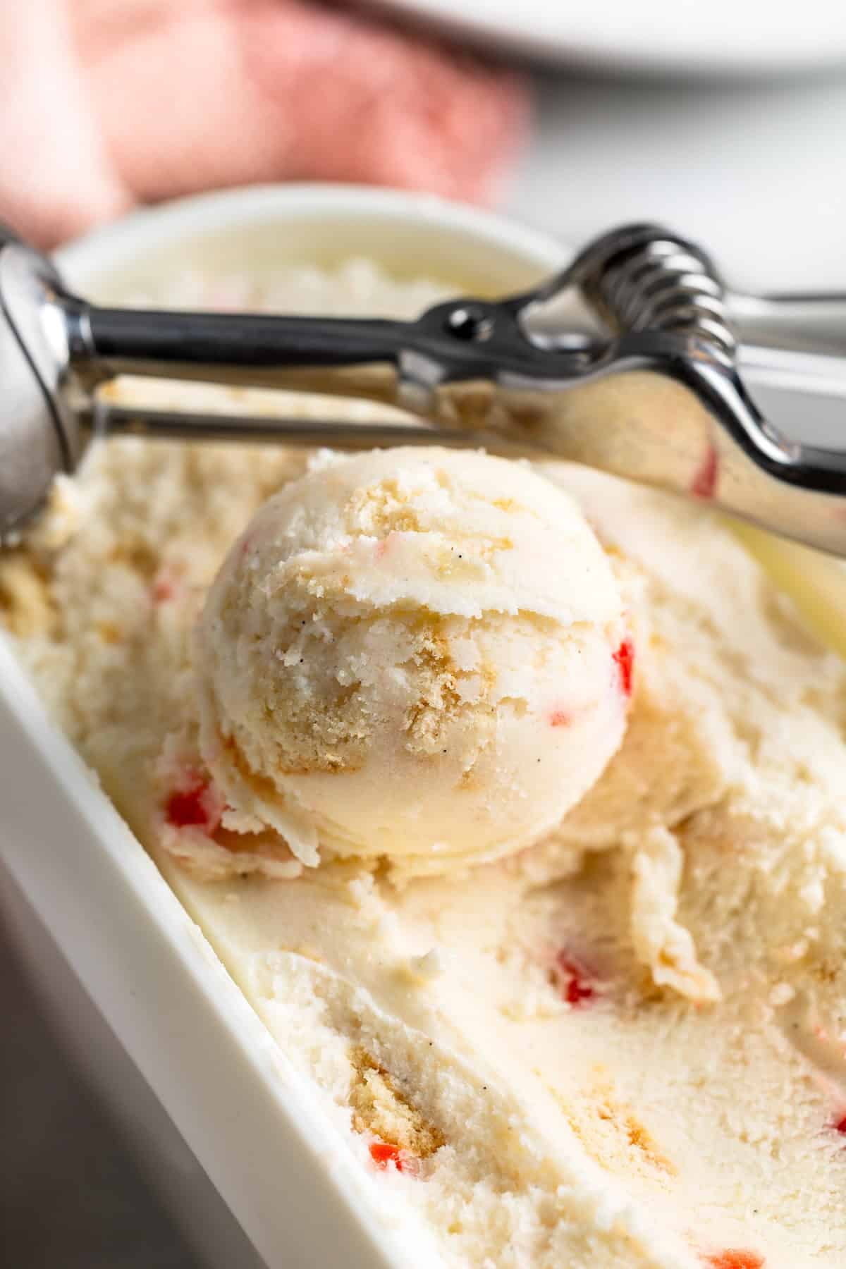 A scoop of pineapple upside down cake frozen yogurt in its tub dotted with vanilla wafer and maraschino cherry bits.