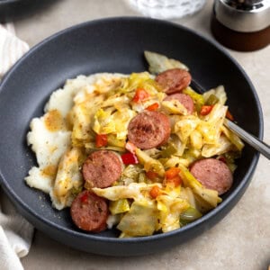 A serving of fried cabbage and sausage over mashed potatoes in a shallow bowl.