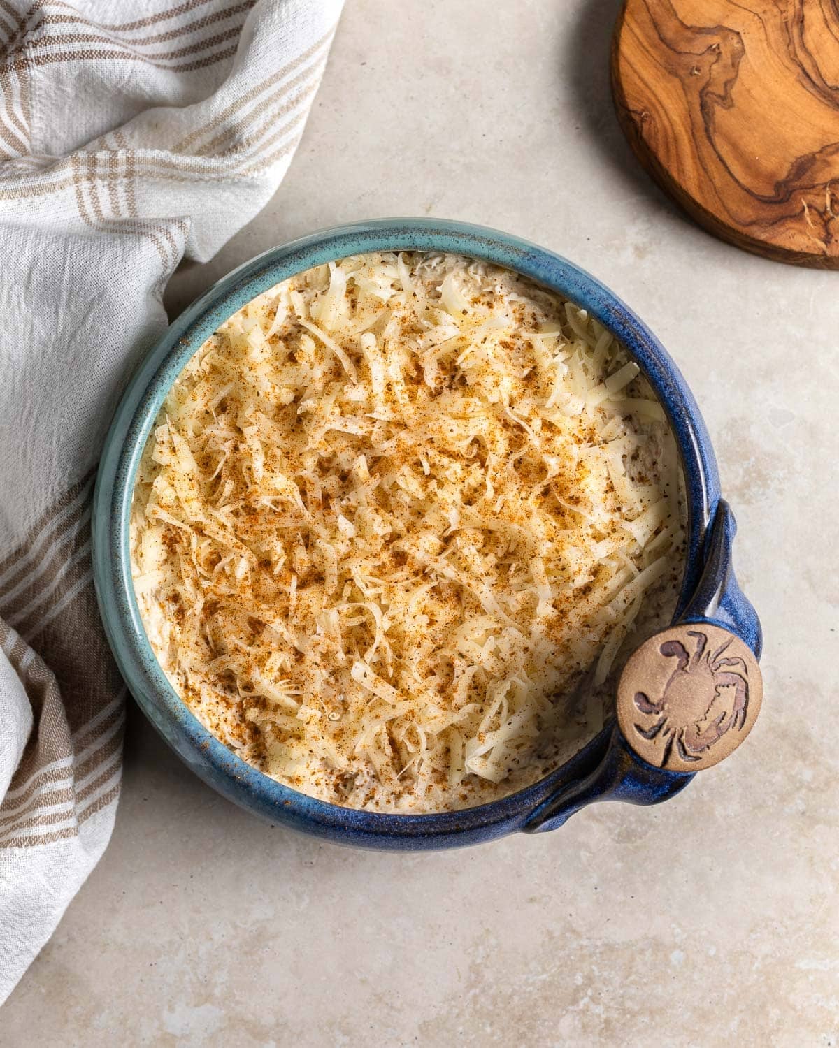 Crab dip in a stoneware bowl pre-oven topped with shredded cheese and sprinkled with Old Bay.