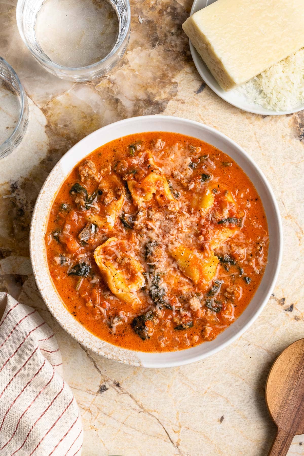 Sausage and tortellini soup in a bowl next to a glass of water and a wedge of Pecorino Romano.