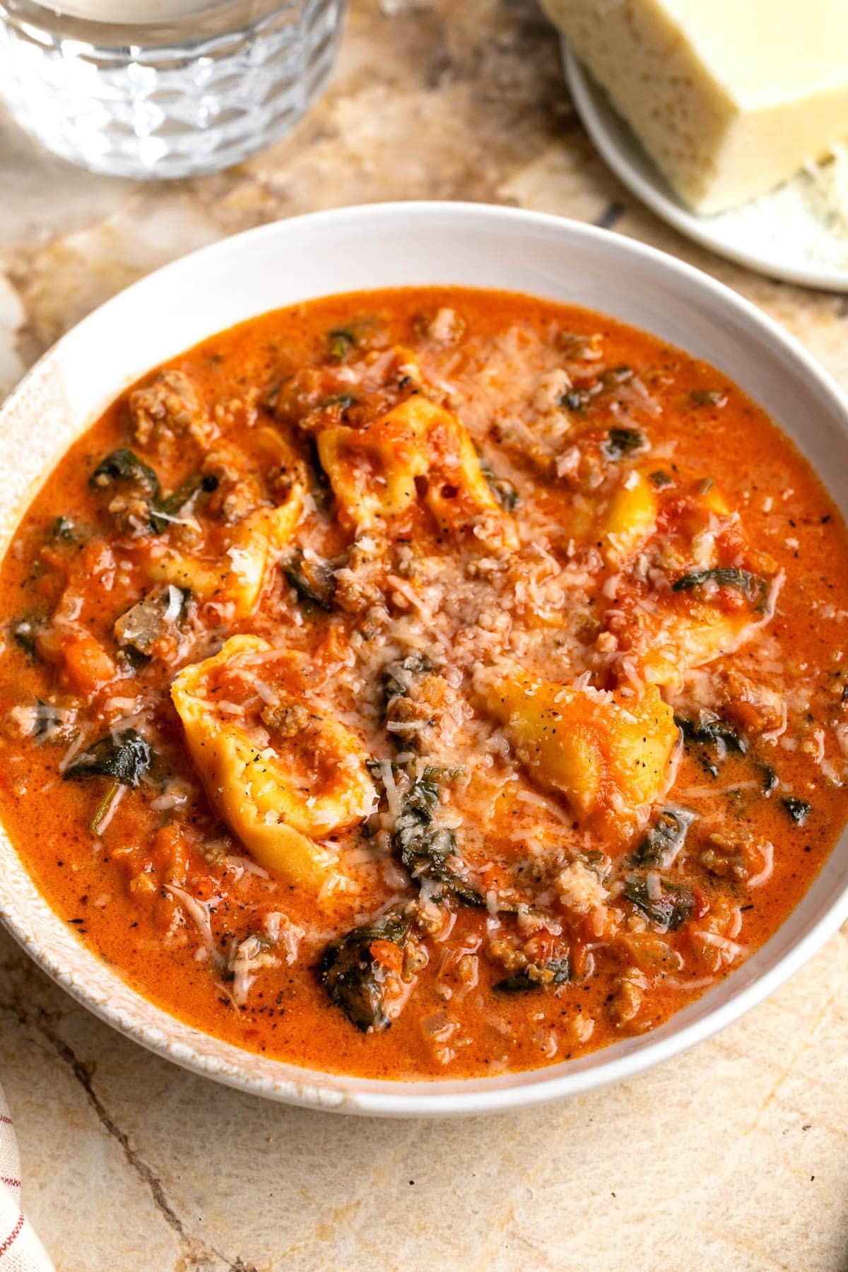Sausage tortellini soup in a bowl next to a wedge of cheese.