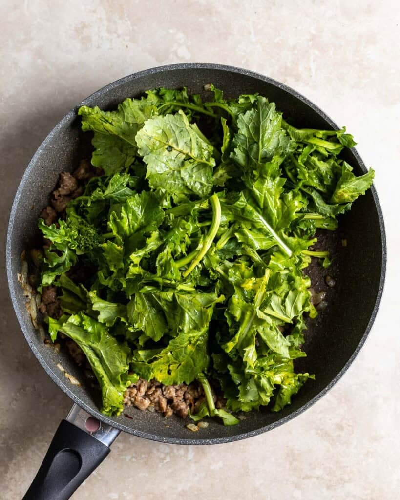 Broccoli rabe leaves being added to the pan of sausage and simmering chicken broth/water.
