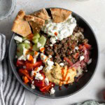 A bowl of dill spiced beef, hummus, brown rice, veggies, feta, and pita.