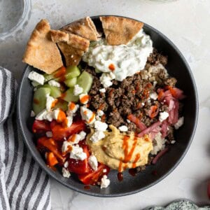 A bowl of dill spiced beef, hummus, brown rice, veggies, feta, and pita.