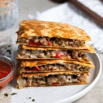 Four quadrants of an Italian sausage quesadilla stacked on top on one another on a plate with a small bowl of dipping marinara.
