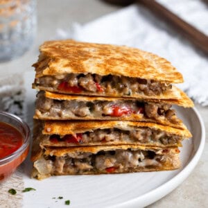 Four quadrants of an Italian sausage quesadilla stacked on top on one another on a plate with a small bowl of dipping marinara.