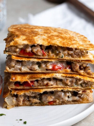 Four quadrants of an Italian sausage quesadilla stacked on top on one another on a plate with a small bowl of dipping marinara.