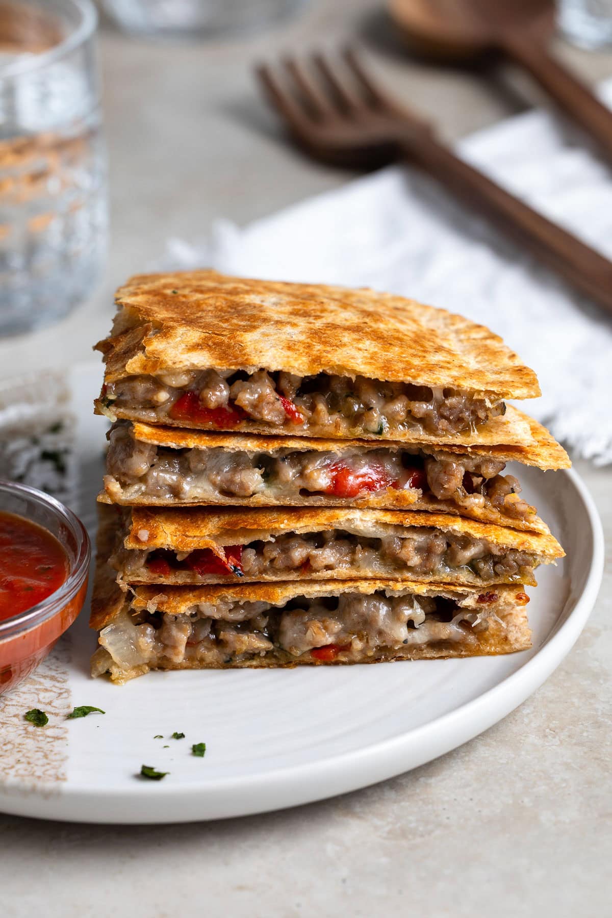 Four quadrants of an Italian sausage quesadilla stacked on top on one another on a plate with a small bowl of dipping marinara.