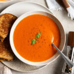 A shallow bowl of tomato soup made with canned tomatoes on a plate with a grilled cheese sandwich accompaniment.