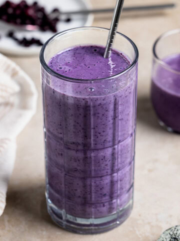 A wild blueberry smoothie in a tall glass with a metal straw and a short glass.