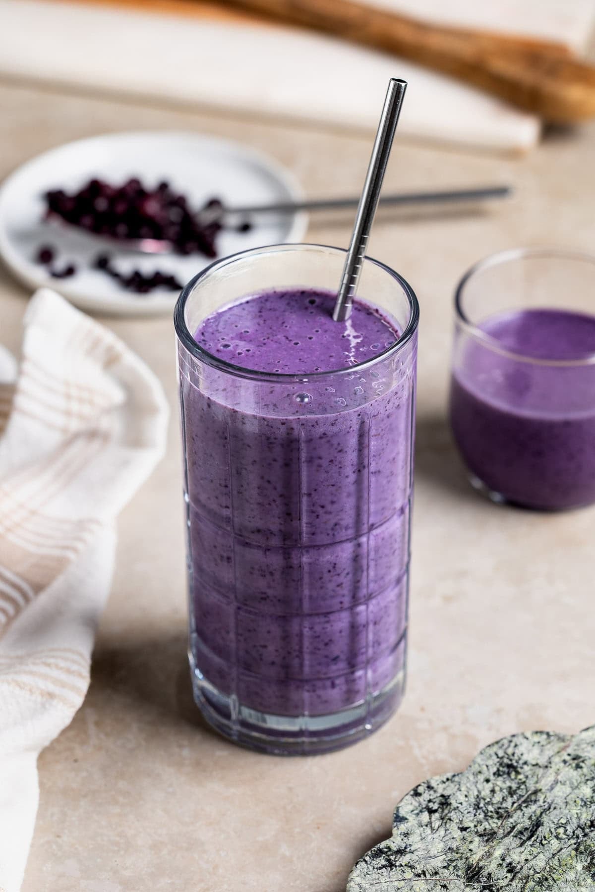 A wild blueberry smoothie in a tall glass with a metal straw and a short glass.