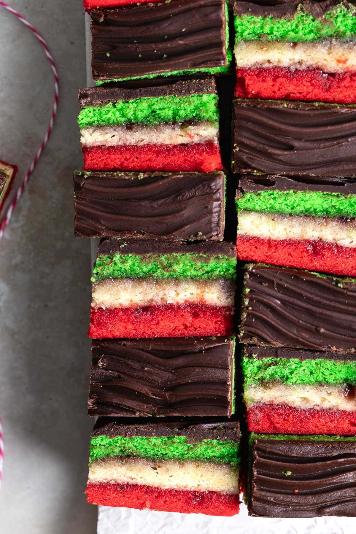 A batch of rainbow cookies situated next to one another in an alternating pattern showing the side layers and wavy chocolate tops.