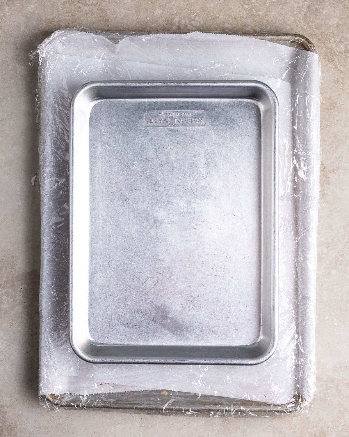 An assembled slab of rainbow cookies wrapped in parchment paper and plastic wrap with a pan on top of it.